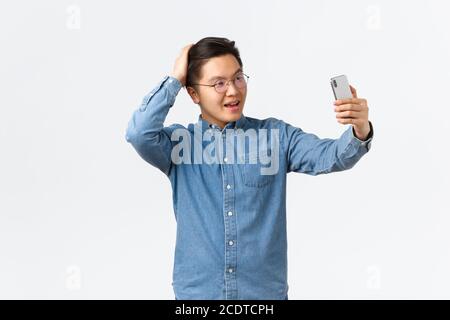 Cheeky confident asian man in glasses and braces feeling sassy, taking selfie, brushing hair with hand, posing for photo, using filter app on mobile Stock Photo