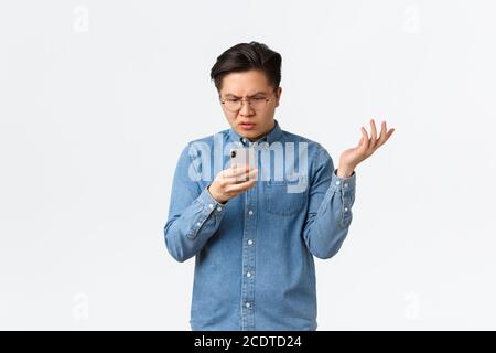 Frustrated and confused asian guy in glasses looking puzzled at mobile phone screen, raising hand up questioned, cant understand what happening online Stock Photo