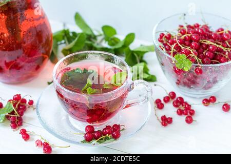 Redcurrant drink in transparent glass carafe and cup. Clear glass vase with red currant berries on the white wooden background. Stock Photo