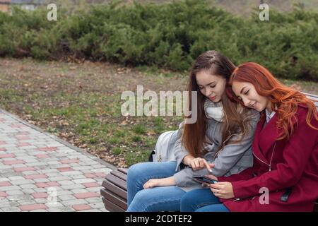 Female teenagers listening to music on smartphone sitting on the bench in an autumn city park Stock Photo