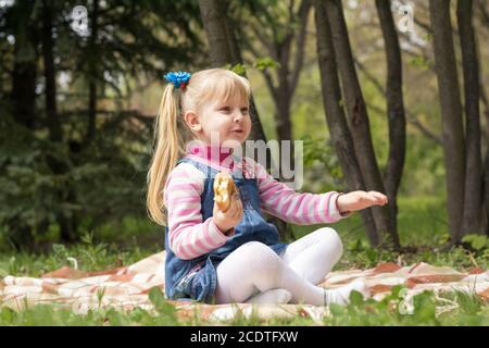 Cute little blonde girl with two ponytails relaxing with a book and a bun in the city park on a spring sunny day. Stock Photo