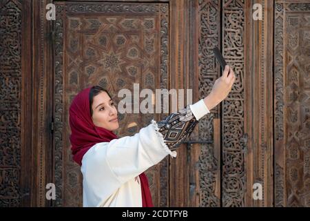 Young muslim woman taking selfie with mobile phone in traditional clothing with red headscarf Stock Photo