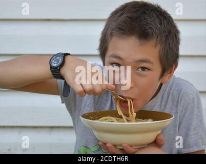 Happy mixed race pre-teen boy (East Asian and Caucasian) eats dry noodles with a fork from a ceramic bowl. Stock Photo