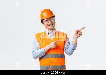 Building sector and industrial workers concept. Cheerful smiling asian builder, construction manager in helmet and reflective clothing pointing upper Stock Photo