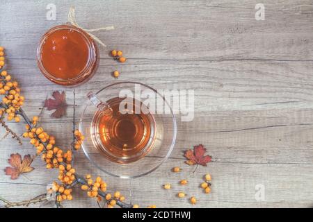 Autumn healthy hot drink concept. Branch of common sea buckthorn with berry, cup of tea, jar of jam on light wooden background. Stock Photo