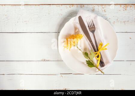 White empty plate, yellow chrysanthemum flowers, napkin, fork and knife tied with a yellow ribbon on light wooden background. Stock Photo