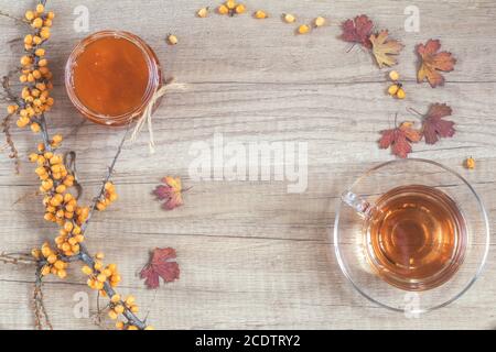 Autumn healthy hot drink concept. Branch of common sea buckthorn with berry, cup of tea, jar of jam on light wooden background Stock Photo