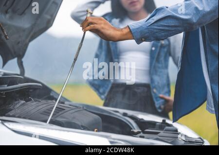 Asian man open car hood for repair as maintenance service with woman background between long road trip. Car broken and breakdown in mountain meadow wi Stock Photo