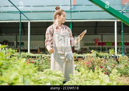 Young blond female farmer in workwear looking at photo of plant in touchscreen Stock Photo
