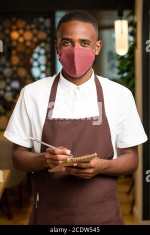 Happy young African man in brown apron and protective mask taking orders Stock Photo