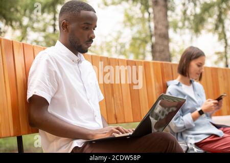Serious guy of African ethnicity in casualwear networking on bench in park Stock Photo