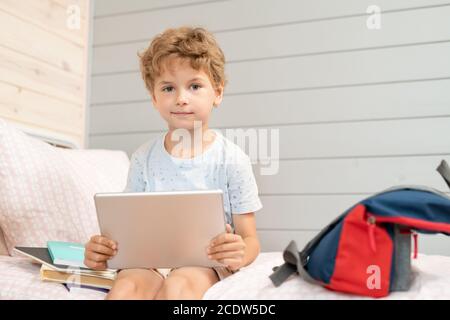 Adorable blond schoolboy with digital tablet sitting on bed after school Stock Photo