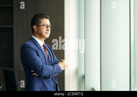 portrait of a asian business leader senior manager standing by window thinking Stock Photo