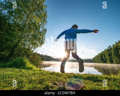 Crazy boy is making fun and jumping on lake bank against evening sunset. Funny childhood. Stock Photo