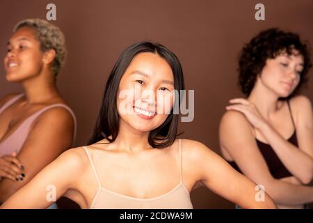 Young cheerful Asian woman in beige tanktop standing in front of other females Stock Photo