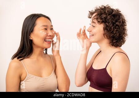 Two young laughing intercultural females in tanktops applying rejuvenating cream Stock Photo