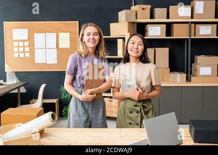 Two young cheerful female workers of online shop office standing by table Stock Photo