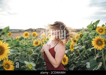 Carefree young woman tossing her long blond hair in sunflower field against sky Stock Photo