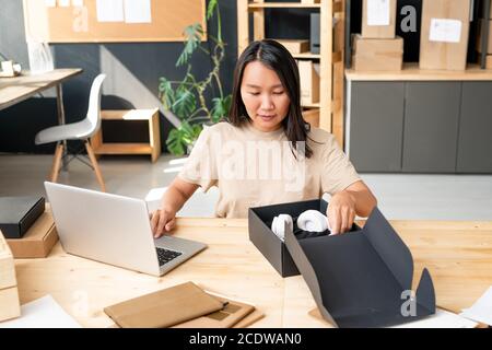 Young Asian female manager of online shop office putting headphones into box Stock Photo
