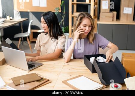 Young blond female manager of online shop office talking to clients on phone Stock Photo
