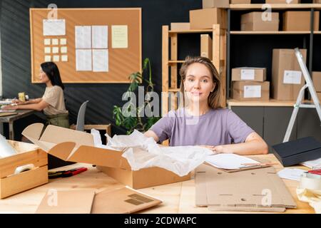 Young female worker of online shop office sitting by table in front of camera Stock Photo