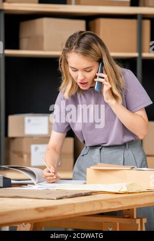 Young woman calling one of clients while checking information about order Stock Photo