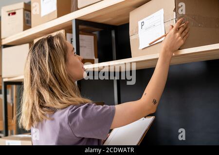 Young blond woman with list of orders and pencil checking address of receiver Stock Photo