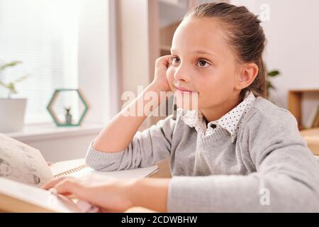 Smiling clever schoolgirl with open book looking through questions on blackboard Stock Photo