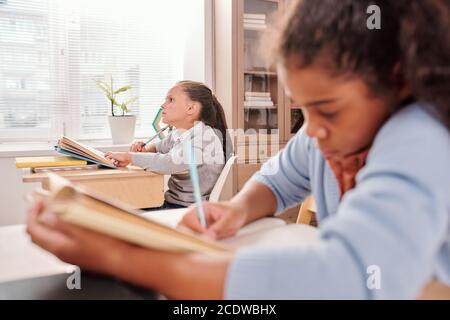 Pensive schoolgirl with open book thinking of answer to question on blackboard Stock Photo