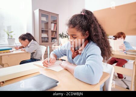 Serious mixed-race schoolgirl making notes or wtiting essay in copybook Stock Photo