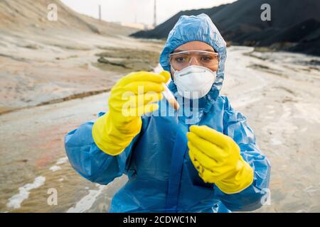 Young female ecologist in respirator and eyeglasses looking at flask with water Stock Photo