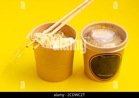 Chicken or vegetable broth with noodle and egg in craft containers on yellow paper background. Soup to go, healthy food delivery. Stock Photo