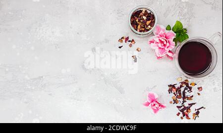 Composition with hot hibiscus tea in glass mug with pink flowers and dry tea leaves on light grey concrete background. Stock Photo