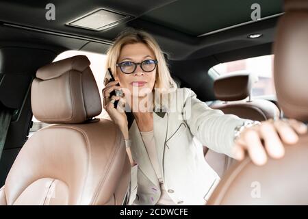 Mature elegant female with blond hair talking on smartphone on backseat of car Stock Photo