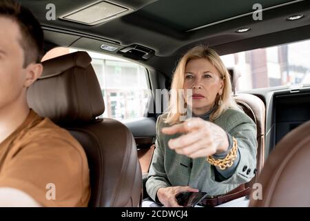 Mature blond businesswoman sitting on backseat of taxi cab and pointing on road Stock Photo