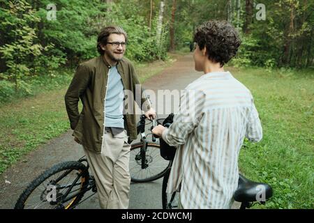Young casual couple with bicycles standing on road between trees and talking Stock Photo