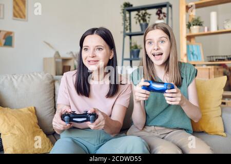 Excited teenage girl with joystick sitting on couch next to her happy mother