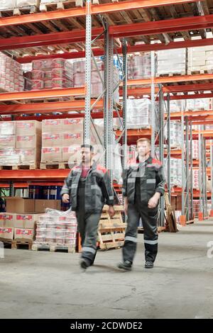Two young intercultural male workers in uniform moving along large factory Stock Photo