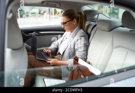 Working in taxi. Side view of a successful business woman wearing eyeglasses using laptop while sitting on back seat in the car Stock Photo