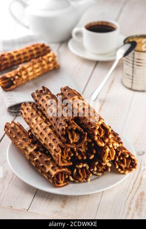 Still life composition, waffle tubules with creamy and milk jam, coffee cup on white wooden background, food and homemade baking concept in rustic sty