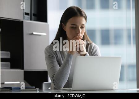 Thoughtful focused young businesswoman looking at laptop screen in office Stock Photo