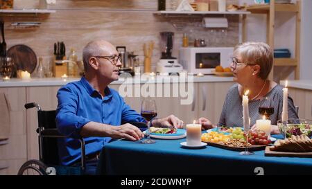 Aged senior man with disabilities having romantic dinner sitting at the table in cozy kitchen. Wheelchair immobilized paralyzed handicapped man dining with wife at home, enjoying the meal Stock Photo