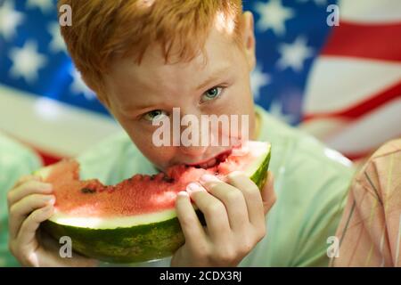 Close up portrait of freckled red-haired boy holding watermelon and looking at camera with American flag in background, copy space Stock Photo