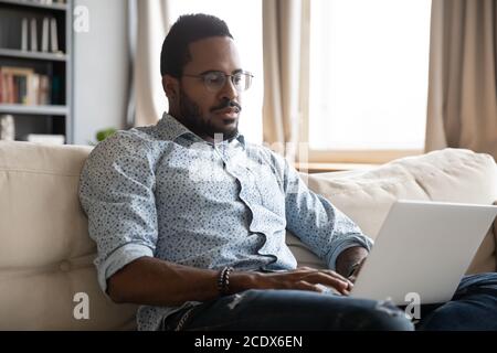 Serious focused African American man working on laptop on couch Stock Photo