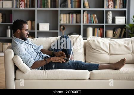 Positive African American man lying on couch, using laptop Stock Photo