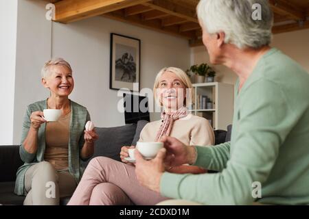 Two smiling senior women drinking tea and talking to man while sitting on sofa in the living room Stock Photo