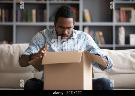 Unhappy shocked African American man unboxing parcel, looking inside box Stock Photo