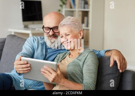 Happy senior couple sitting on sofa and watching movie on digital tablet at home Stock Photo