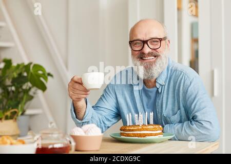 Portrait of senior bearded man in eyeglasses sitting at the table with birthday cake drinking tea and smiling at camera Stock Photo