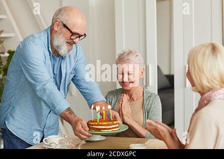 Happy senior woman sitting at the table while her friends bringing the birthday cake and congratulating her Stock Photo